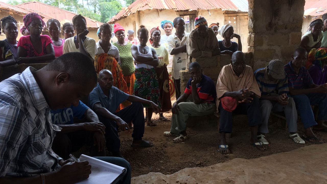 Meeting with men and women gathered, one man sitting and taking notes some other sitting while majority stand in the background