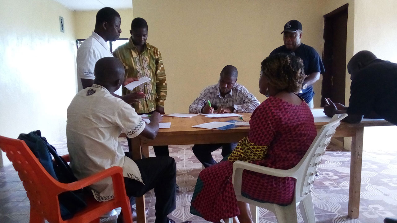 Small group of 6 men and 1 woman standing and sitting around a table, one male taking notes