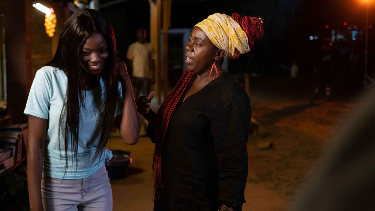 Nighttime scene with two women, one is smiling and looking down and the other is talking to her