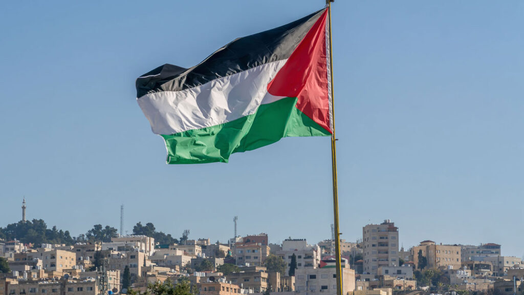 The large Palestinian flag is waiving above the city with tall buildings in the background