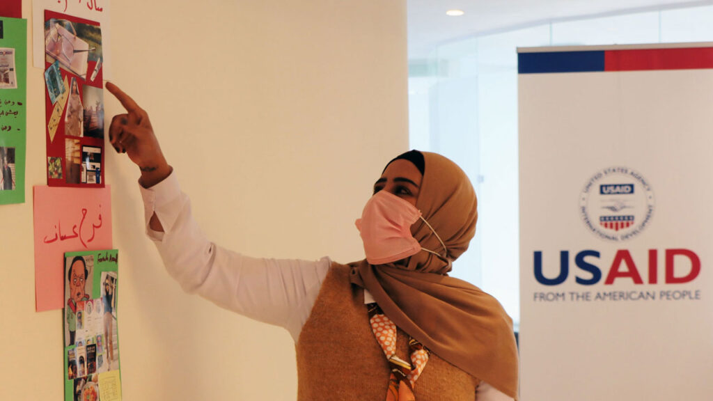 Woman wearing facemask pointing at collage with USAID banner in the background