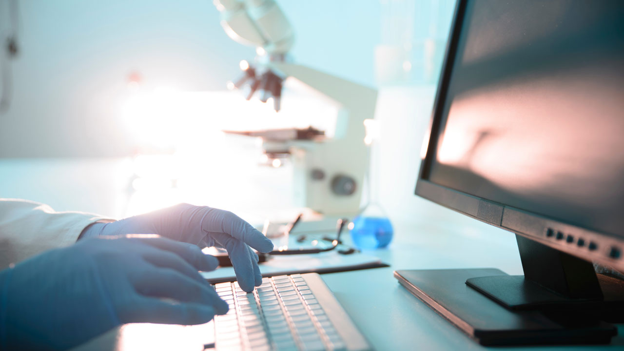 Gloved hands typing on computer keyboard with microscope in the background