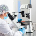 A person in a white lab coat is looking through a microscope