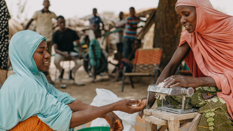 Two women feature one making food, the other with one hand stretch to collect food and the other holding coins